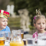 Zwei kleine Kinder mit Kronen sitzen an einem Tisch im Freien mit Getränken und Speisen, darunter Orangensaft und Kuchen, in einem Garten.