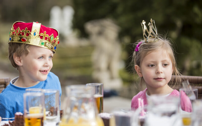 Zwei kleine Kinder mit Kronen sitzen an einem Tisch im Freien mit Getränken und Speisen, darunter Orangensaft und Kuchen, in einem Garten.