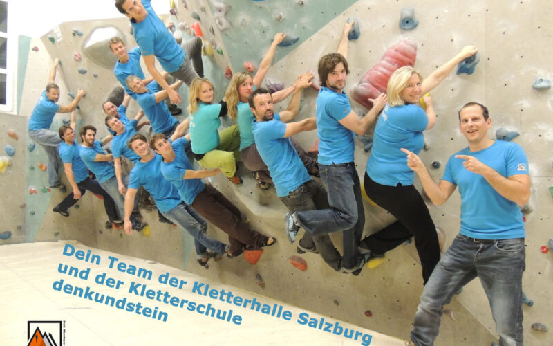 Eine Gruppe von Personen in passenden blauen Hemden posiert an einer Indoor-Kletterwand in der Kletterhalle Salzburg, mit Text und Logos für die Kletterhalle und Kletterschule am unteren Rand.