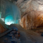 Große Eisformation in einer Felsenhöhle, die von blauem Licht beleuchtet wird, mit einem Weg und verstreuten Steinen auf dem Höhlenboden.
