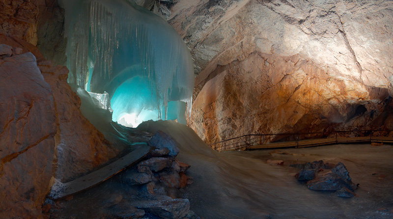 Große Eisformation in einer Felsenhöhle, die von blauem Licht beleuchtet wird, mit einem Weg und verstreuten Steinen auf dem Höhlenboden.