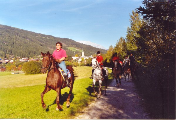 Vier Personen reiten auf einem unbefestigten Weg neben einem grasbewachsenen Feld, mit einem Dorf und bewaldeten Hügeln im Hintergrund unter einem klaren Himmel.