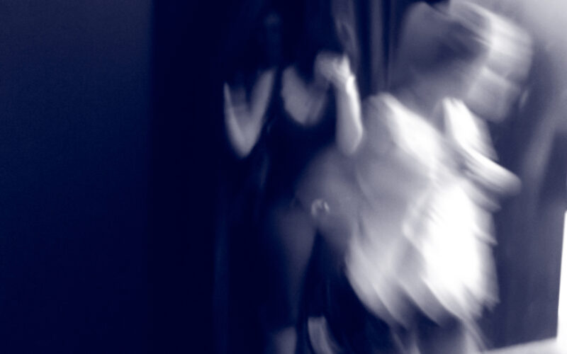 Ein unscharfes Foto zeigt drei Personen in Bewegung, von denen eine durch einen dunklen Vorhang austritt und zwei andere vorbeigehen; unten links erscheint der Schriftzug "S I N N E".