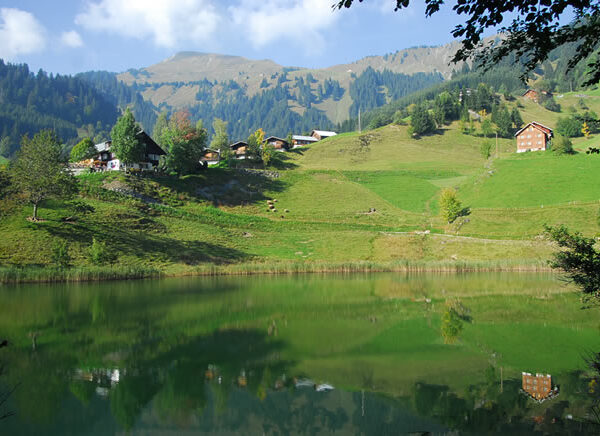 In einem ruhigen See spiegeln sich grüne Hügel, verstreute Häuser und bewaldete Berge unter einem teilweise bewölkten Himmel.