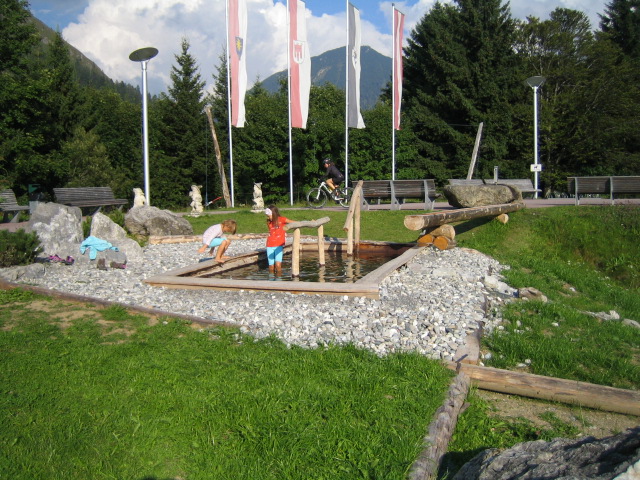 Zwei Kinder spielen in einem flachen, felsigen Wasserspiel in einem Park mit Bänken, Fahnen und einer Kulisse aus Bäumen und Bergen.