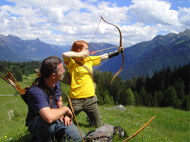 Ein Kind spannt einen Bogen und zielt einen Pfeil, während ein Erwachsener zuschaut. Beide befinden sich im Freien auf einem grasbewachsenen Hügel mit Bergen und Bäumen im Hintergrund unter einem teilweise bewölkten Himmel.