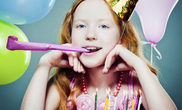 Ein junges Mädchen mit langen roten Haaren trägt einen Partyhut und hält eine Partytorte im Mund. Vor ihr stehen Luftballons und eine Geburtstagstorte mit brennenden Kerzen.