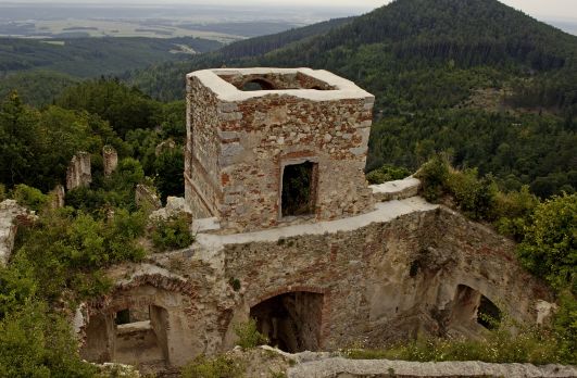Ruinen eines alten Steingebäudes mit einem quadratischen Turm, umgeben von Grün und bewaldeten Hügeln im Hintergrund.