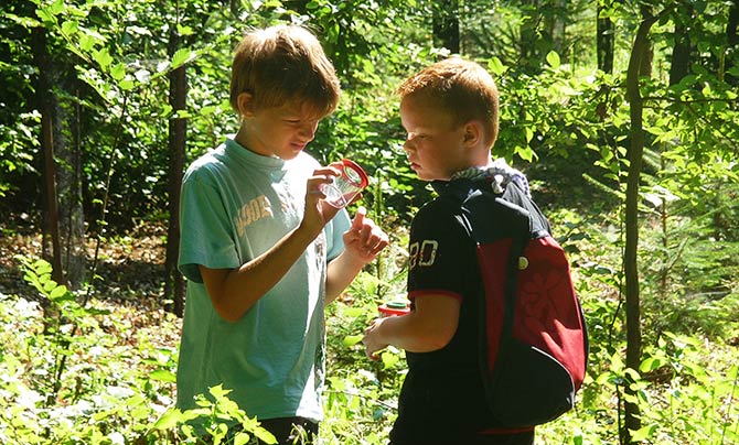 Zwei Jungen stehen in einem sonnenbeschienenen Wald. Der eine hält eine Lupe in der Hand und untersucht einen Gegenstand, während der andere, der einen Rucksack trägt, zusieht.