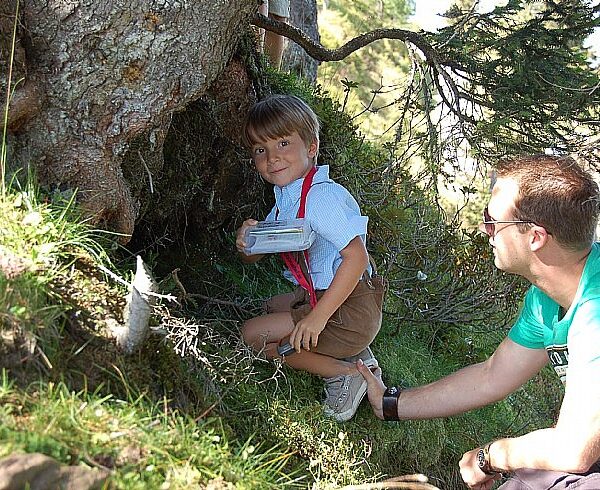 Ein kleiner Junge kauert an einem Baum und hält einen Behälter in der Hand, während ein erwachsener Mann mit Sonnenbrille neben ihm auf einer Wiese kniet.