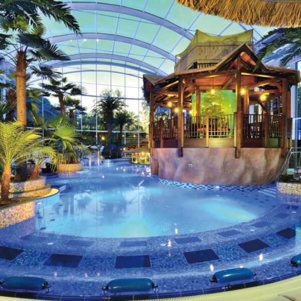 Tropischer Innenpool mit Palmen, blauen Kacheln und einem zentralen Holzpavillon unter einer großen Glaskuppel.