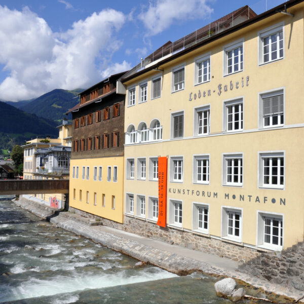 Ein gelbes Gebäude mit der Aufschrift "Kunstforum Montafon" steht neben einem fließenden Fluss, mit Bergen und einem Steg im Hintergrund unter einem teilweise bewölkten Himmel.