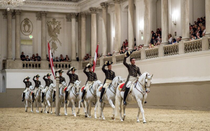 Eine Gruppe von uniformierten Reitern auf weißen Pferden salutiert in Formation in einer verzierten Reithalle vor den Augen der Zuschauer.