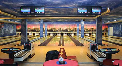 Eine hell beleuchtete Bowlingbahn mit mehreren Bahnen, Bildschirmen für die Punktevergabe, Sitzgelegenheiten und einem dekorativen Wandgemälde an der Rückwand.