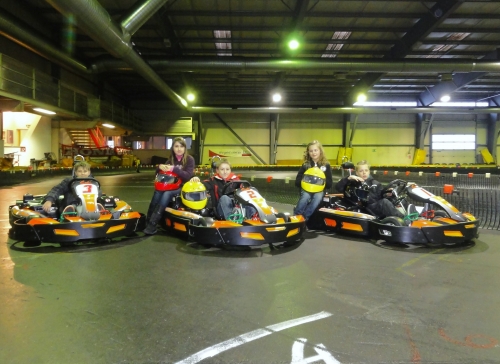 Fünf Kinder sitzen in Gokarts, die nebeneinander auf einer Indoor-Gokartbahn aufgereiht sind, tragen Helme und Rennanzüge und sind bereit zum Fahren.