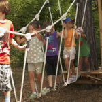 Mehrere Kinder laufen über eine Seilbrücke auf einem von Bäumen umgebenen Holzspielplatz.