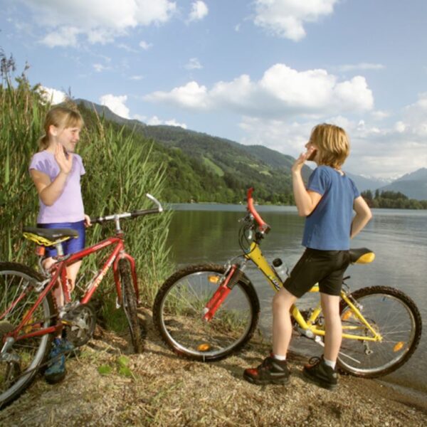 Zwei Kinder mit Fahrrädern stehen an einem Seeufer, umgeben von Grün und Bergen unter einem teilweise bewölkten Himmel.