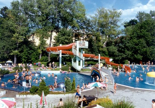 Öffentliches Freibad mit einer großen Wasserrutsche, umgeben von Bäumen und Menschen, die an sonnigen Tagen schwimmen, spielen und sich sonnen.