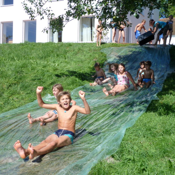 Kinder rutschen eine selbstgebaute Wasserrutsche auf einem Grashügel hinunter, oben wartet eine Gruppe und im Hintergrund ist ein weißes Gebäude zu sehen.