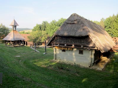 Ein reetgedecktes Haus und ein hölzerner Glockenturm stehen auf einer Wiese, die von Bäumen und einem Holzzaun umgeben ist.