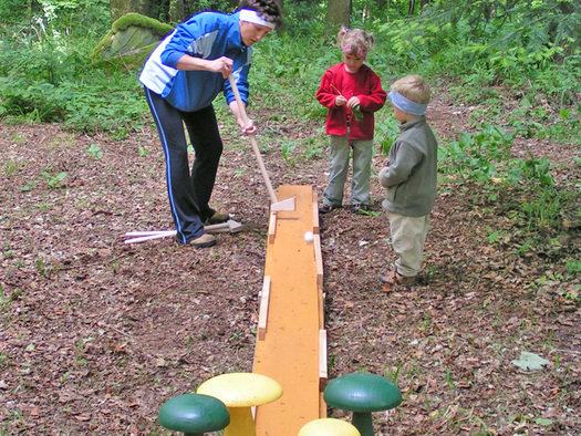 Ein Erwachsener und zwei Kinder spielen im Freien ein Spiel, bei dem sie mit Holzschlägern einen Ball über einen schmalen, erhöhten Weg in einem Waldgebiet schlagen.