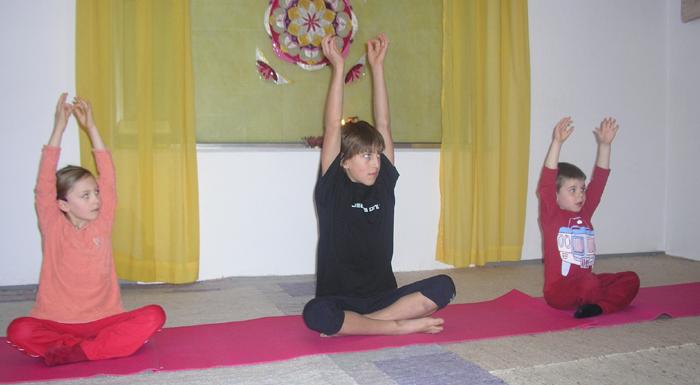Drei Kinder sitzen im Schneidersitz auf Yogamatten in einem Haus und heben ihre Arme über den Kopf. Im Hintergrund sind gelbe Vorhänge und ein dekorativer Wandbehang zu sehen.