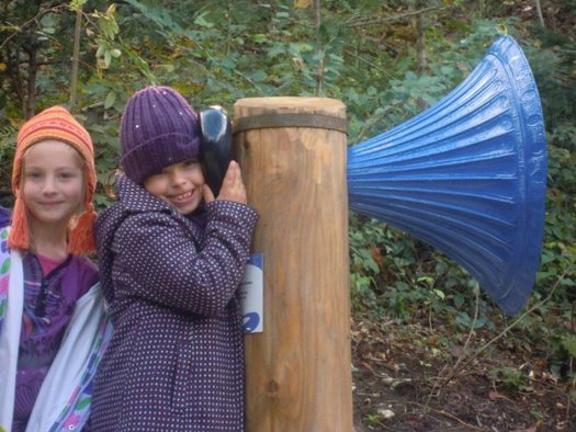 Zwei Kinder stehen an einem Holzpfosten, an dem ein großes blaues Megaphon befestigt ist; ein Kind hört am Pfosten zu, während das andere neben ihr im Freien steht.