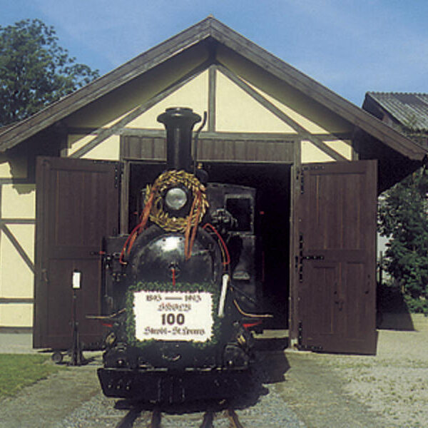 Eine mit einem Kranz geschmückte alte Dampflokomotive steht vor einem offenen Holzschuppen unter einem strahlend blauen Himmel.