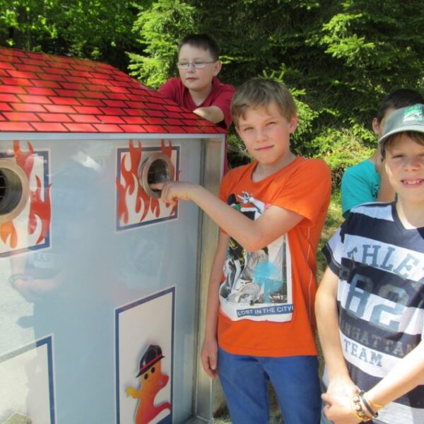 Mehrere Jungen stehen neben einem Modell zur Demonstration des Brandschutzes in Form eines Hauses, wobei ein Junge seine Hand in eine kreisförmige Öffnung an der Seite steckt.