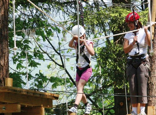 Ein Kind und ein Erwachsener, die Helme und Klettergurte tragen, navigieren an einem sonnigen Tag im Freien zwischen Bäumen durch einen Seilgarten.