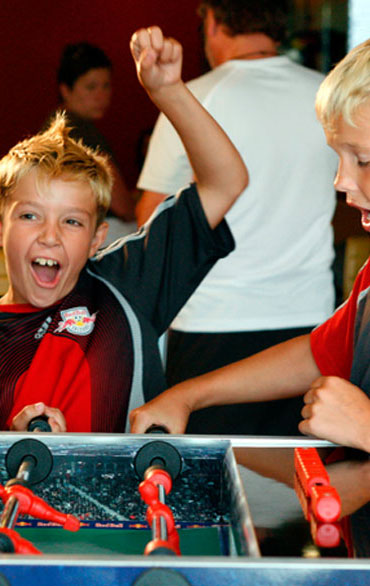 Zwei Jungen spielen Tischfußball; der eine jubelt mit erhobener Faust und aufgeregtem Gesichtsausdruck, während der andere auf den Tisch blickt.