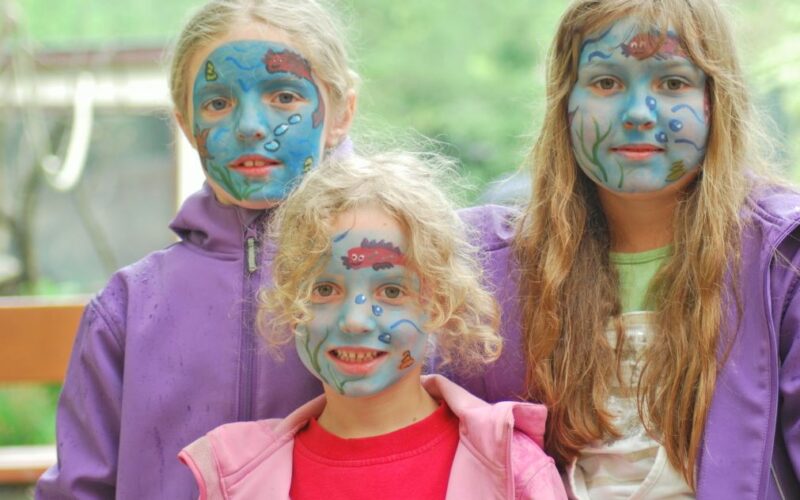 Drei Kinder mit blauer Gesichtsbemalung und Unterwassermotiven stehen zusammen im Freien, alle tragen lila oder rosa Jacken.