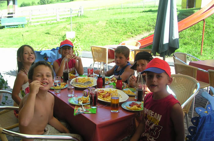 Sechs Kinder sitzen um einen Tisch im Freien, essen Pommes frites und trinken Limonade, während im Hintergrund eine Spielplatzrutsche und eine Wiese zu sehen sind.