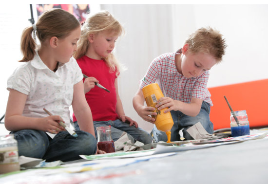 Drei Kinder sitzen auf dem Boden und malen; eines gießt gelbe Farbe, während die anderen Pinsel halten. Um sie herum sind Malutensilien und Farbdosen verstreut.