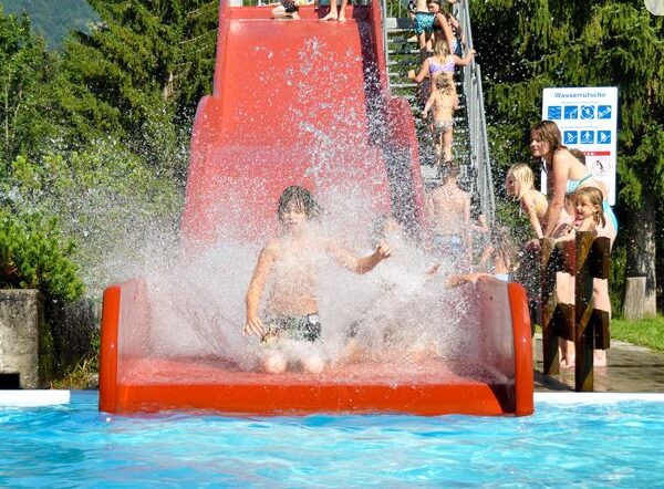 Ein Kind spritzt in ein Becken am Ende einer roten Wasserrutsche, während andere Kinder in der Schlange warten und Bäume im Hintergrund zu sehen sind.