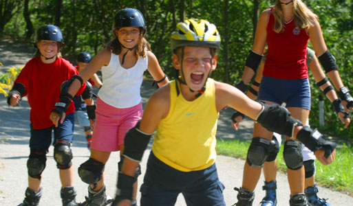 Fünf Kinder mit Helmen und Schutzschuhen fahren gemeinsam auf einem gepflasterten Weg im Freien, umgeben von Grün.