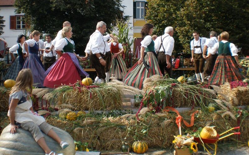 Menschen in traditioneller Kleidung tanzen im Kreis auf einer mit Stroh bedeckten Plattform; ein Mädchen sitzt auf einem großen Kürbis im Vordergrund, umgeben von Herbstdekorationen.