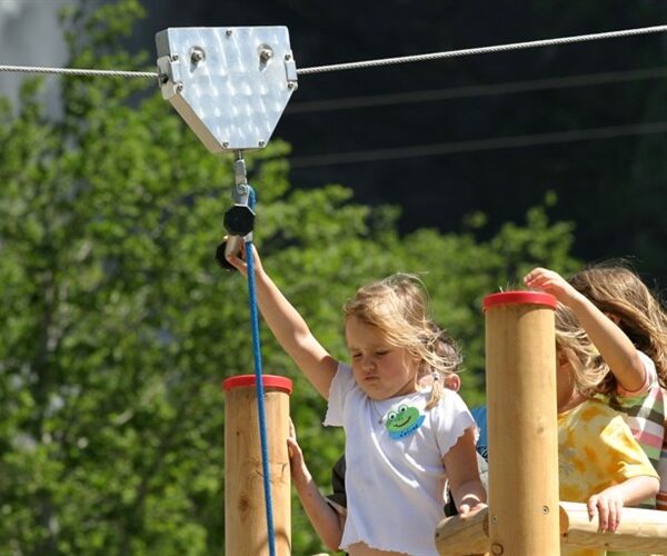 Mehrere Kinder warten auf einer hölzernen Plattform, während ein Mädchen sich darauf vorbereitet, an einem sonnigen Tag im Freien eine Seilrutsche zu benutzen.