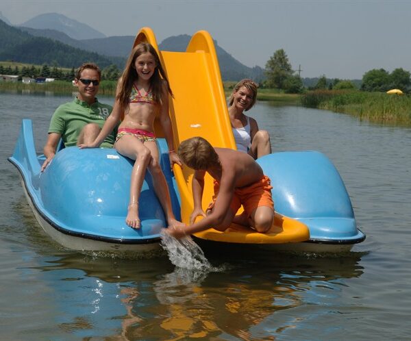 Eine vierköpfige Familie sitzt in einem blauen Paddelboot mit gelber Rutsche auf einem See; der Junge vorne greift ins Wasser, während die anderen lächeln.