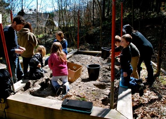 Eine Gruppe von Kindern und Erwachsenen ist an einem sonnigen Tag um ein Hochbeet versammelt und gräbt und erforscht den Boden.