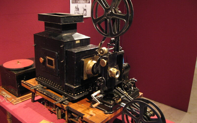 Ein alter Filmprojektor mit großen Rollen und Messingbeschlägen steht auf einem Tisch vor einer roten Wand, mit einem Schwarz-Weiß-Foto im Hintergrund.