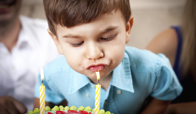 Ein kleiner Junge in einem blauen Hemd bläst zwei brennende Kerzen auf einer Geburtstagstorte aus.
