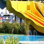 Eine leuchtend gelbe Wasserrutsche führt in einen Außenpool mit bunten Wandmalereien und grünen Bäumen im Hintergrund.