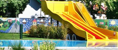 Eine leuchtend gelbe Wasserrutsche führt in einen Außenpool mit bunten Wandmalereien und grünen Bäumen im Hintergrund.