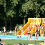 Menschen an einem Freibad mit einer gelben und roten Wasserrutsche, umgeben von Bäumen und Gras, die einen sonnigen Tag genießen.