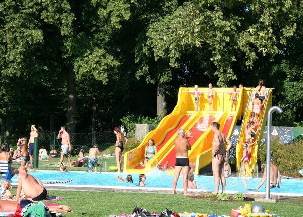 Menschen an einem Freibad mit einer gelben und roten Wasserrutsche, umgeben von Bäumen und Gras, die einen sonnigen Tag genießen.