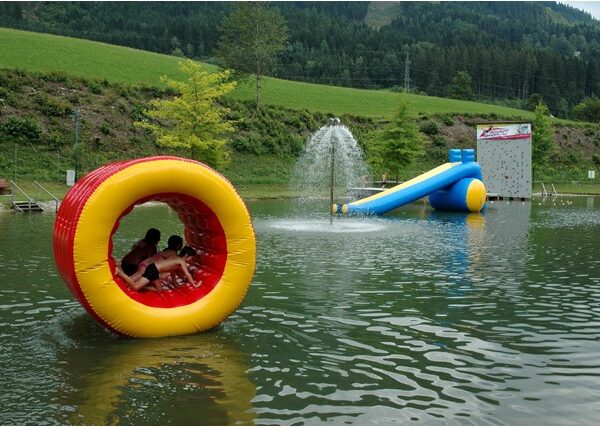 Kinder sitzen in einem großen aufblasbaren gelben und roten Rad, das auf einem Teich schwimmt, während im Hintergrund ein Springbrunnen, eine Wasserrutsche und eine Kletterwand zu sehen sind.