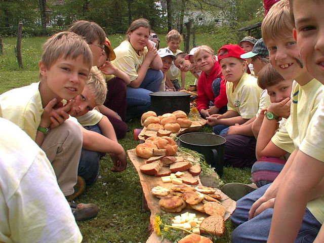 Die Kinder sitzen im Freien auf dem Rasen in zwei Reihen einander gegenüber, und zwischen ihnen liegen Brot, Käse und andere Lebensmittel auf Holzbrettern.