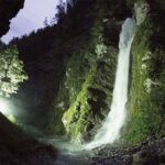 Ein Wasserfall fließt nachts einen von Bäumen umgebenen Felsen hinunter, der von der linken Seite des Bildes aus mit künstlichem Licht beleuchtet wird.