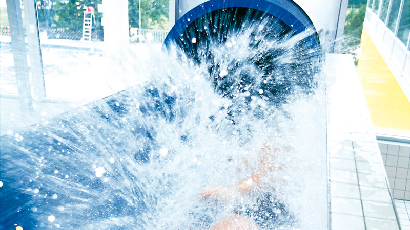 Eine Person verlässt eine Wasserrutsche in einem Innenraum, wobei am Ende der Rutsche ein großer Wasserspritzer entsteht.